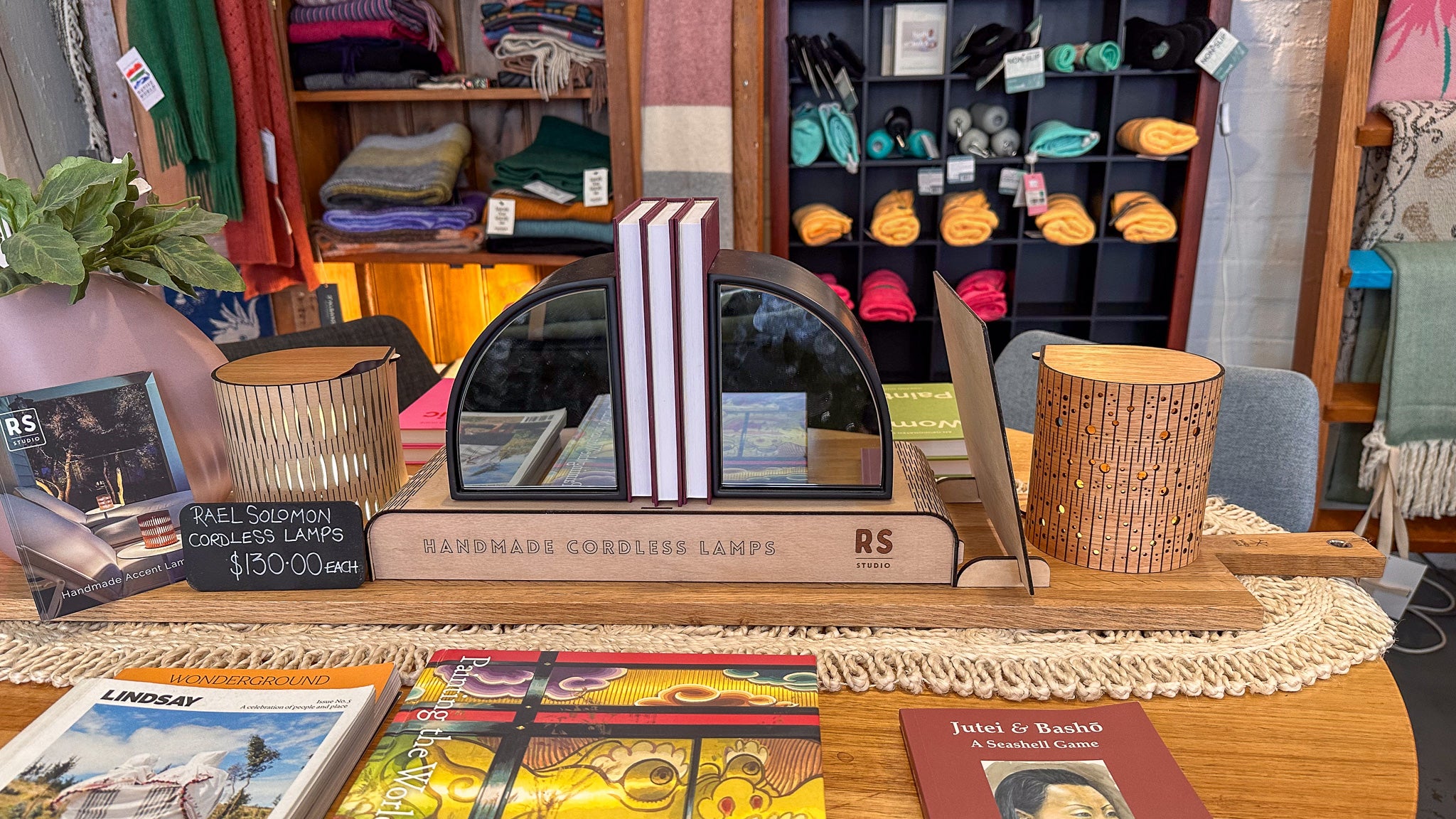 Handmade wooden lampshades with 'RS' branding on a table with books and plants.