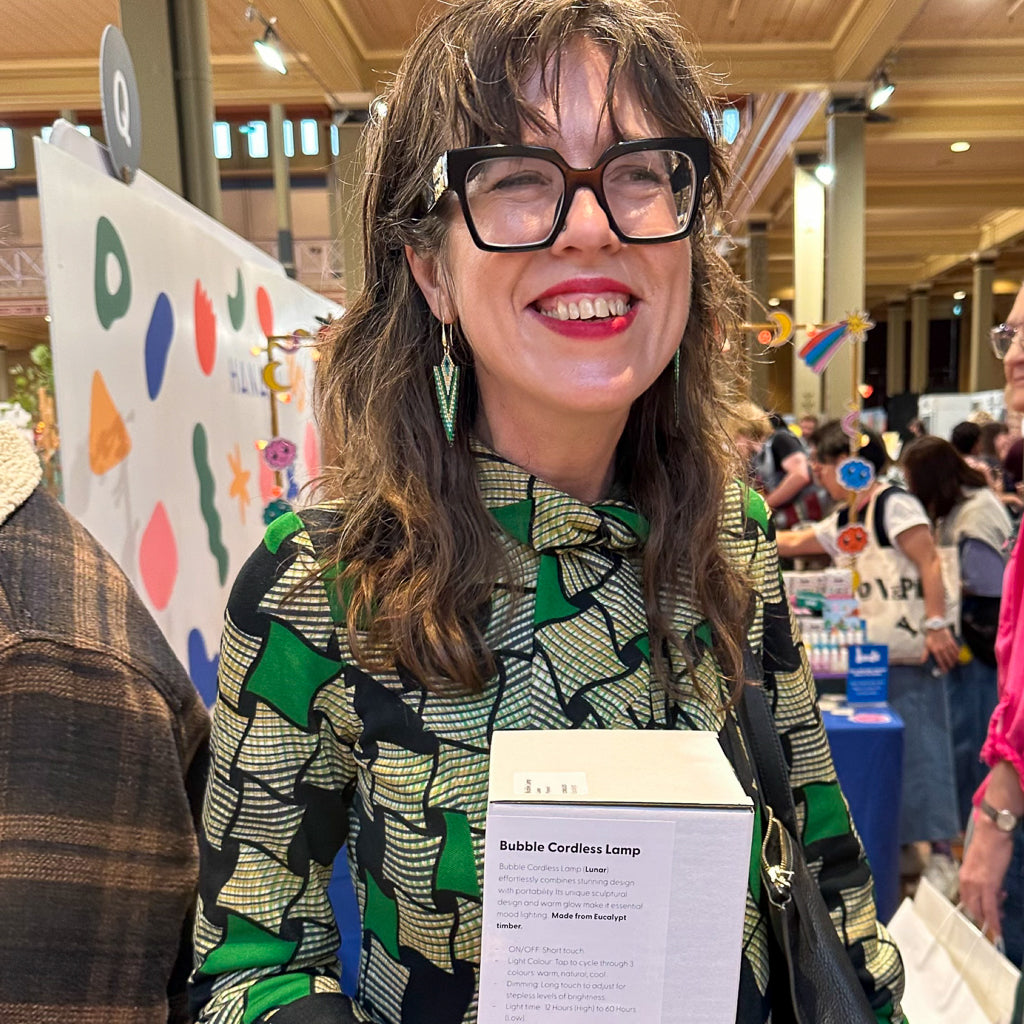 Woman with glasses holding a 'Bubble Cordless Lamp' at a market event with colorful displays and people in the background.